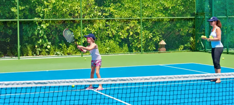 Young girl in cap swings tennis racket on blue court as instructor in cap holds ball nearby, green fence backdrop