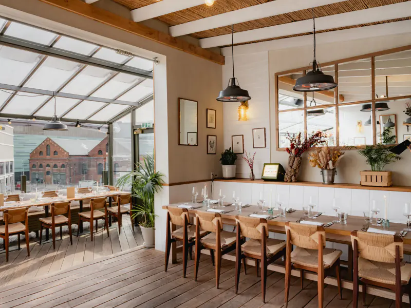 Sophisticated dining room at The Broadcaster, London, with long wooden table set for meal, plants, and panoramic city views through glass doors.