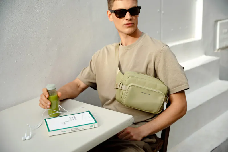 Young man in sunglasses and beige shirt sits at table holding green drink, wearing green Kapten & Son crossbody bag