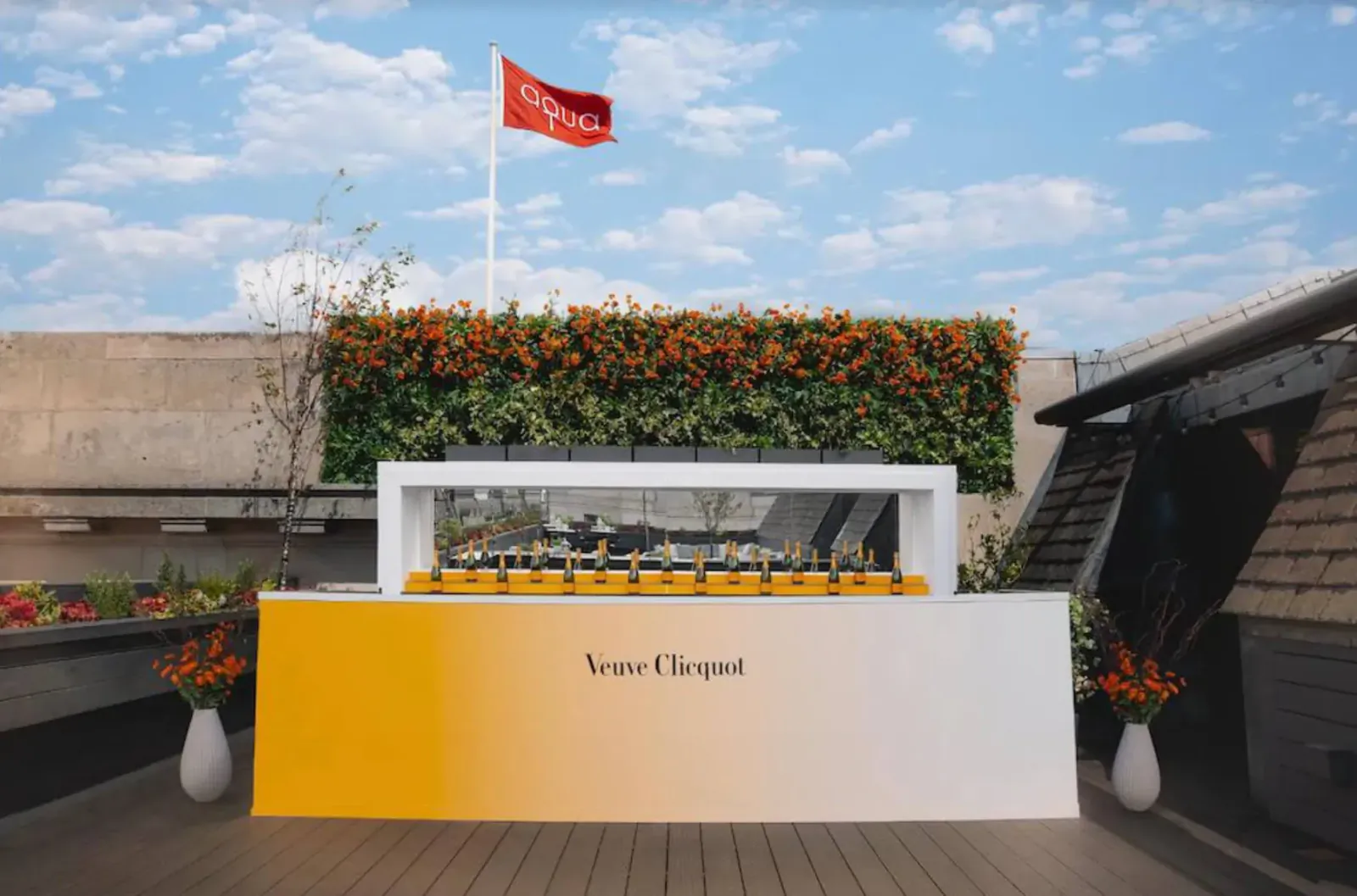 Veuve Clicquot bar on summer terrace with yellow Veuve Clicquot sign, orange flower wall, red flag, and potted plants.