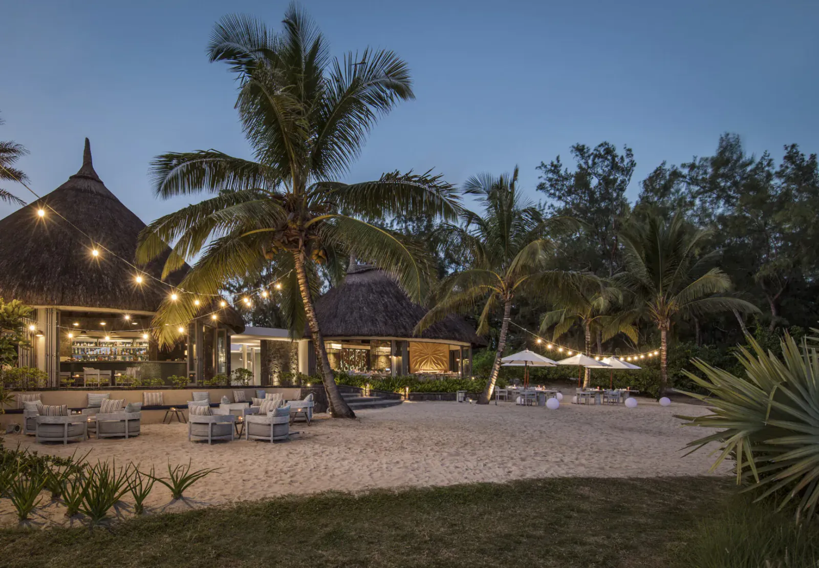 Twilight beachfront resort at Anantara Iko Mauritius with thatched huts, palm trees, string lights, lounge chairs, and umbrellas on sand.