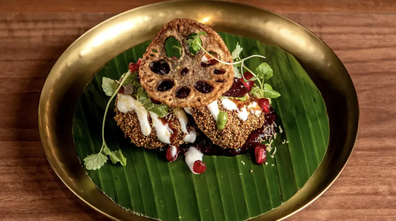 Crispy lotus root fritters topped with yogurt, tamarind sauce, pomegranate, and microgreens on banana leaf in gold bowl