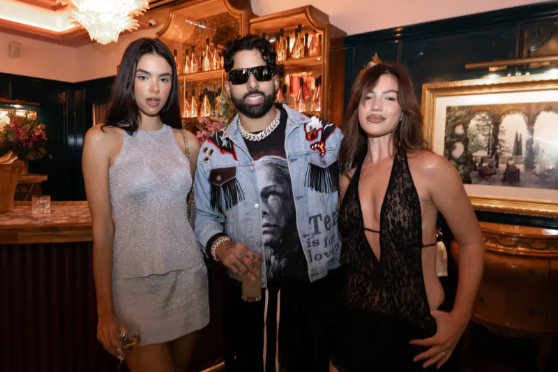 Three stylish Latin artists pose at Armand de Brignac gallery event by wood bar with bottles and chandelier.