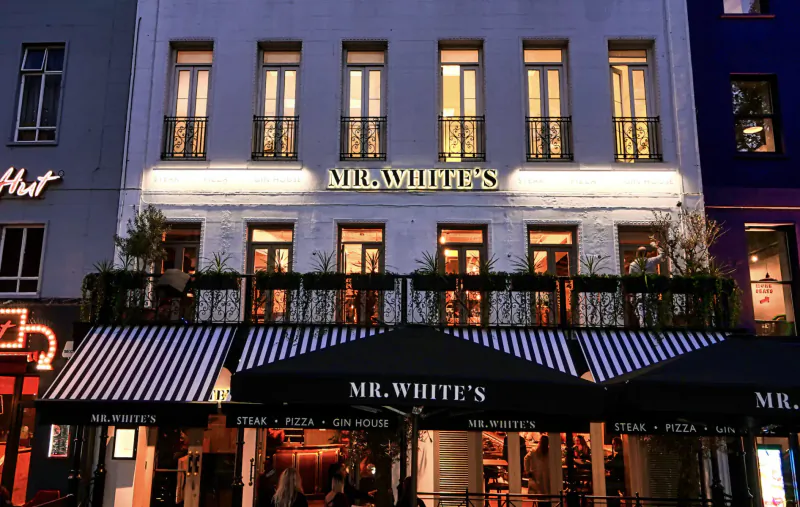 Mr. White's restaurant facade at night in Leicester Square, black-and-white striped awning, neon sign, plants on balcony.