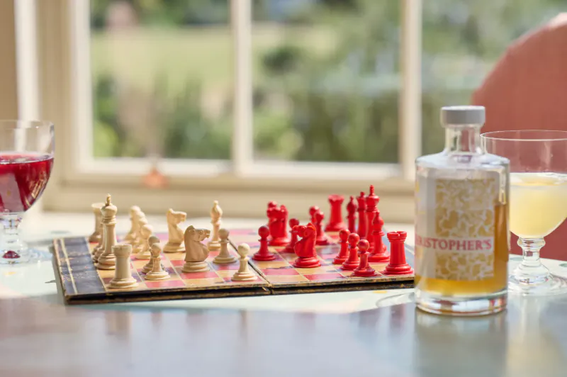 Chessboard with white and red pieces on table beside Christopher's Cordials bottle, two glasses of cordial, window view of fields