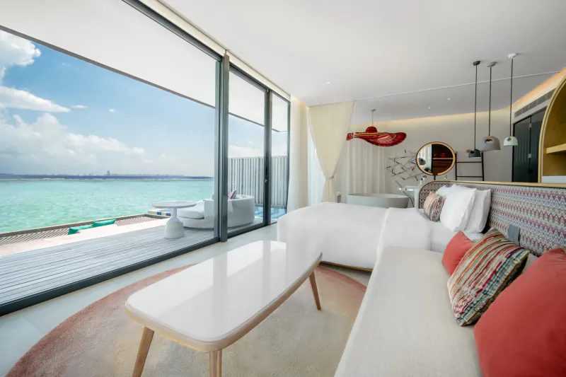 Luxurious SO/ Maldives villa interior with panoramic turquoise lagoon view, white sofa, red pillows, and modern decor.