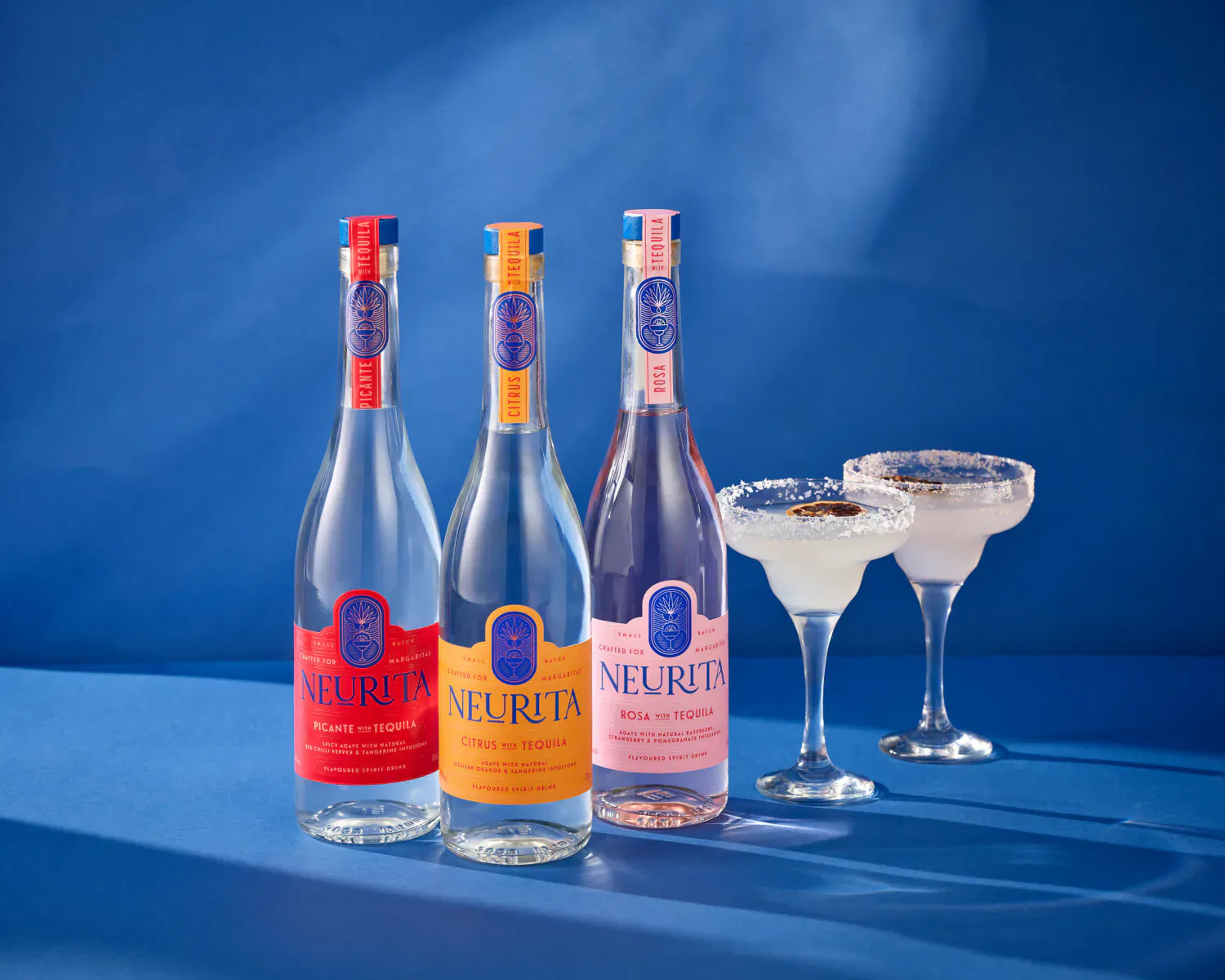 Three Neurita Tequila bottles in red, orange, pink with two margarita glasses on blue background.