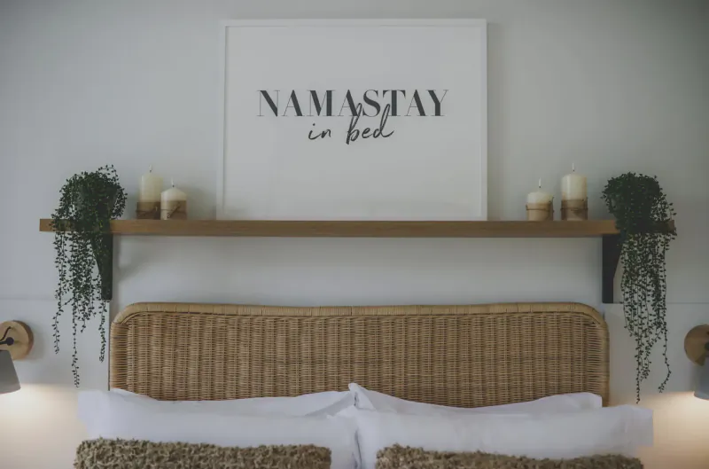 Cozy bedroom with rattan headboard, white pillows, and 'Namastay in bed' wall sign on wooden shelf with plants and candles.