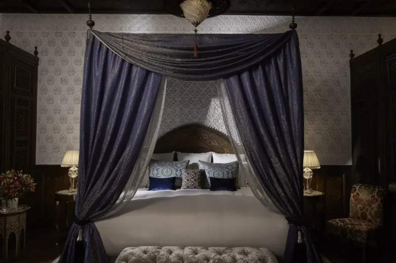 Luxurious four-poster bed with purple canopy in opulent Royal Mansour Marrakech bedroom, ornate lamps and furniture.
