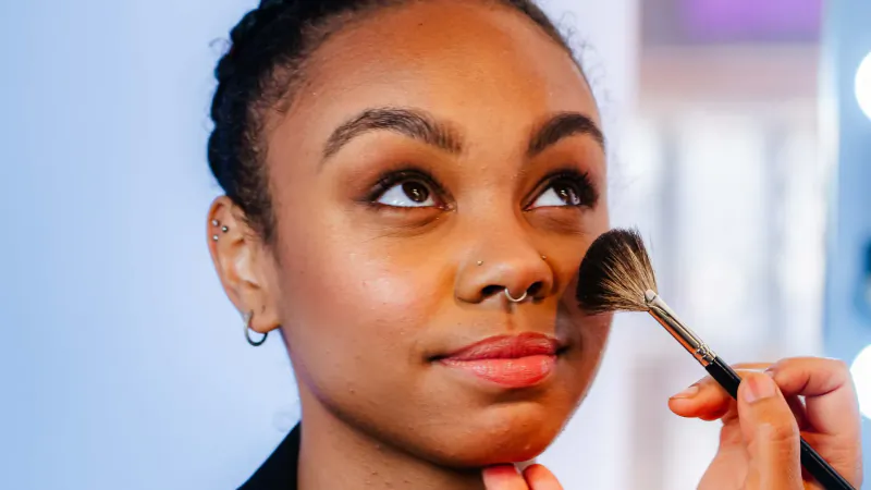 Young Black woman with nose piercing getting makeup applied with brush at Urban Retreat London City salon