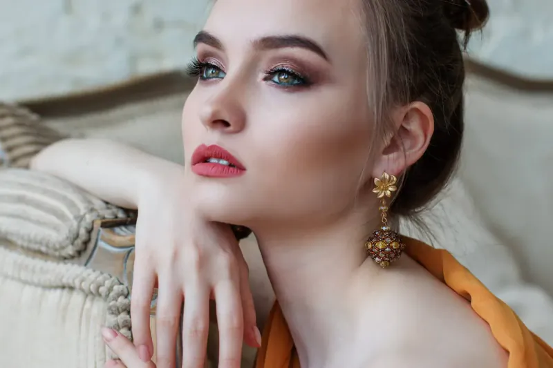Close-up portrait of young woman in Regency-inspired fashion, reclining on ornate sofa with hand on cheek, red lips, gold earrings, orange gown