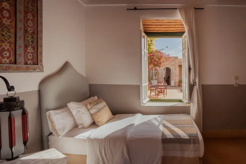 Cozy bedroom in Essaouira riad with curved headboard, lantern, open window to sunny courtyard with red bougainvillea and cafe table.