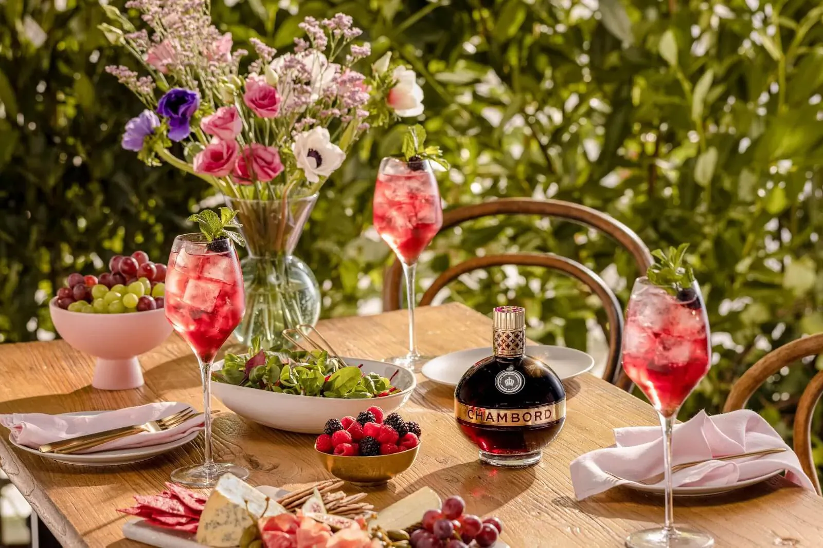 Chambord Berry Spritz: bottle, pink spritz glasses, berries, grapes, cheese board on wooden table amid flowers