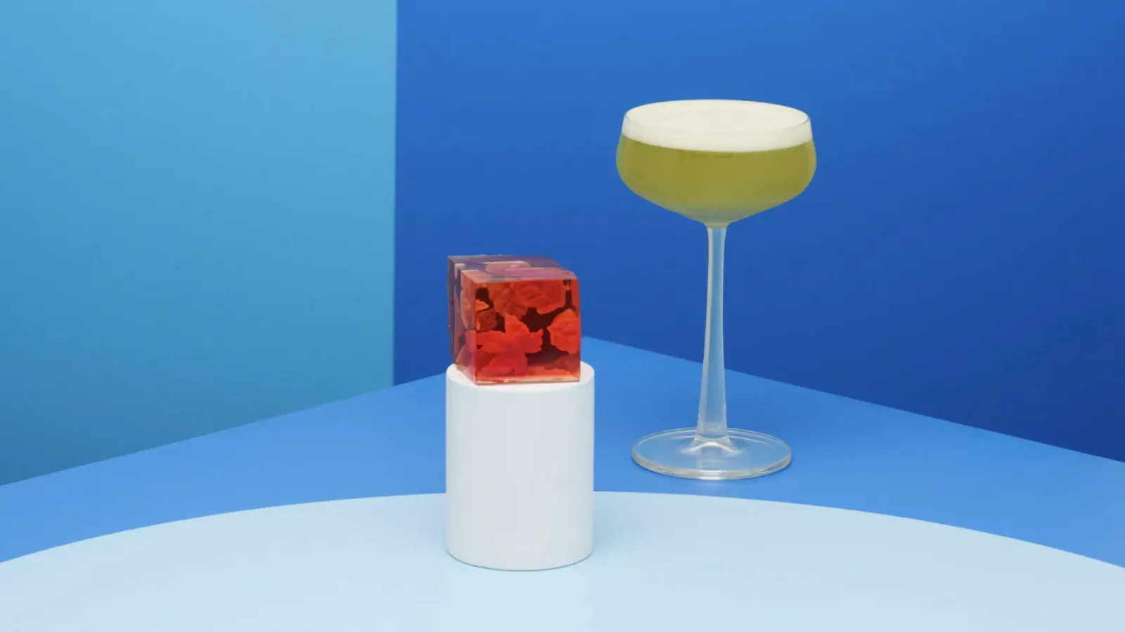 Red cube with devil figure on white base beside green foamy cocktail on blue surface