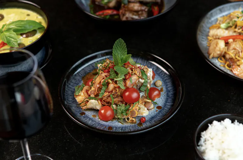 Assortment of Thai dishes: papaya salad with mint and tomatoes, green curry, stir-fry, noodles, rice, and black pitcher on black table.