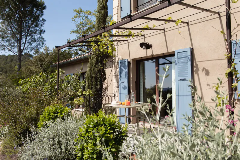 Provencal house exterior with blue shutters, vine-covered pergola, small table with oranges, lush gardens, and trees under blue sky.