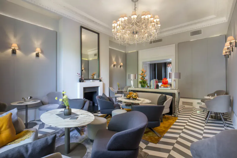 Elegant lounge at Eccleston Square Hotel London with chandelier, blue armchairs, marble tables, flowers, and fireplace.