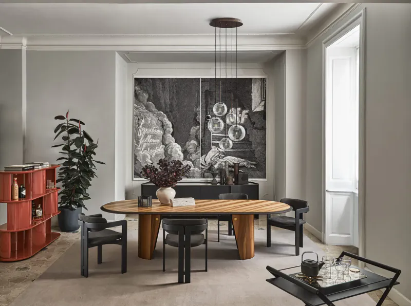 Modern dining room with oval wooden table, black chairs, red bookshelves, potted plants, chandelier, and large abstract wall art.