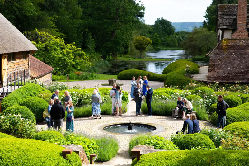 Group of people gathered around fountain in lush gardens with thatched buildings and lake, Nyetimber vineyard visit
