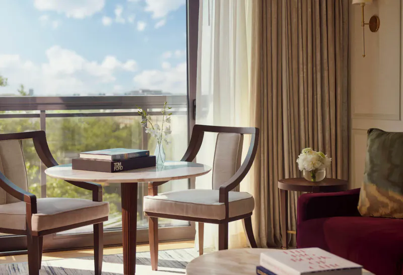 Elegant hotel lounge with round table, upholstered chairs, books, flowers, and balcony view of blue sky and greenery