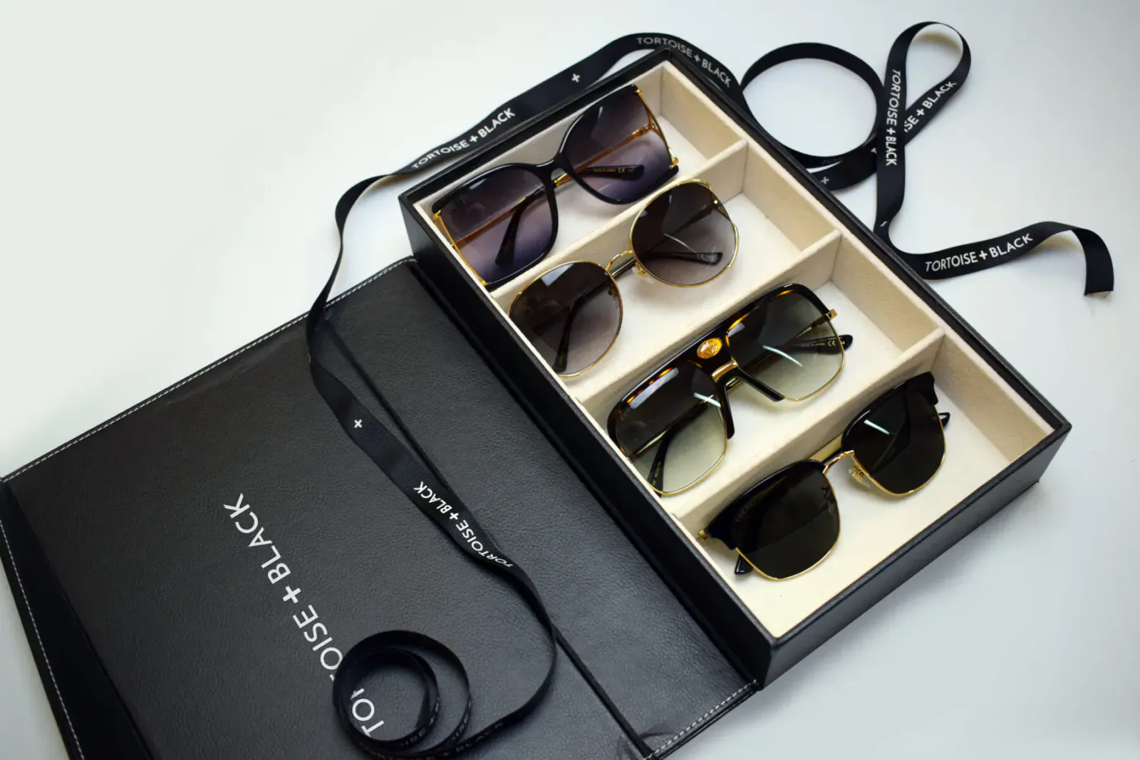 Open black Tortoise+Black luxury eyewear box displaying six pairs of stylish sunglasses in compartments with satin ribbons.