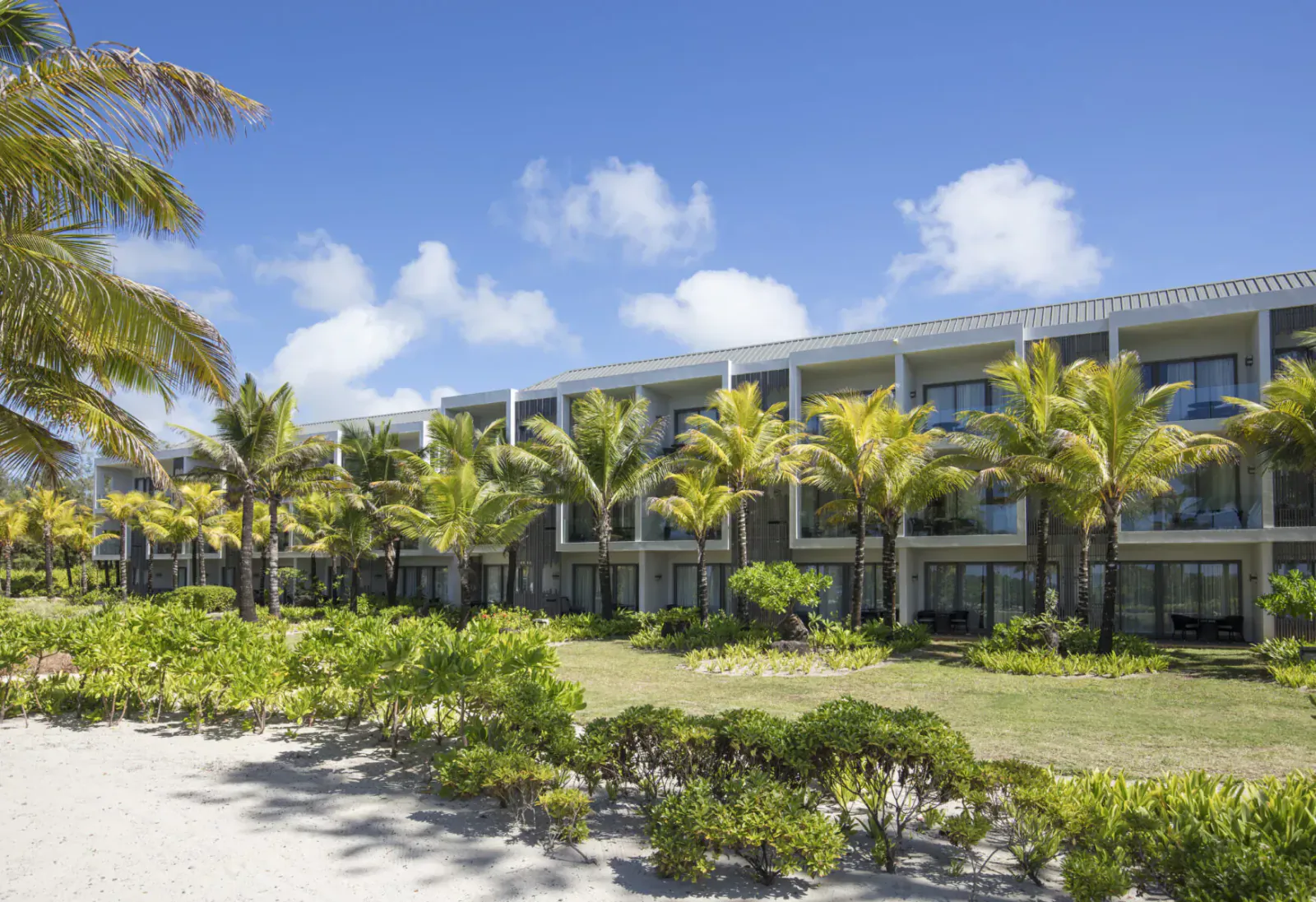 Anantara Iko Mauritius Resort & Villas: modern beachfront hotel with palm trees, lush gardens, and white sand under blue sky.