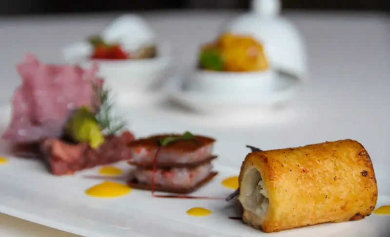 Elegant Michelin-style plate with prosciutto, seared meat, golden stuffed roll, garnishes, and yellow sauce drizzles