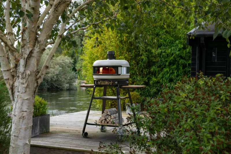 Gozney pizza oven with glowing interior on stand by riverside deck amid trees and shrubs