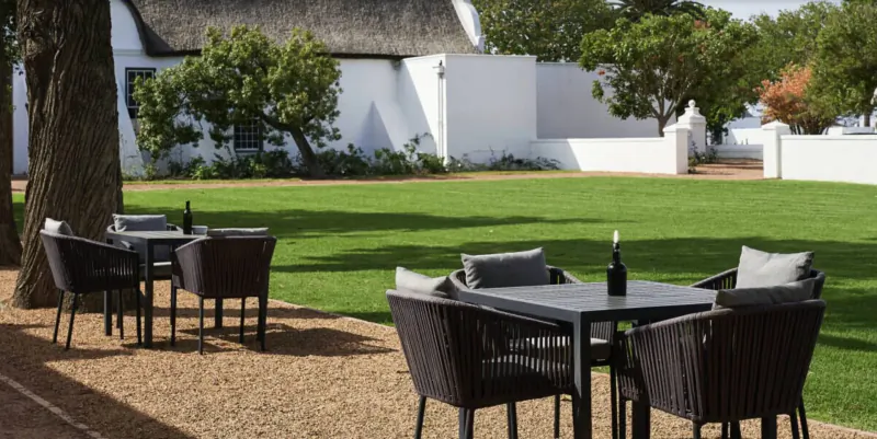 Outdoor seating at Clara’s Barn on Vergenoegd Löw wine estate, with table, wine bottle, cushions, white thatched building, trees, and lawn