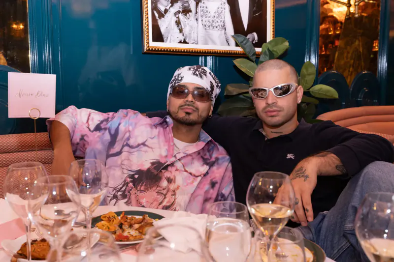 Two bald men in sunglasses at gallery dinner table with wine glasses, pasta, and 'Gala' sign amid teal decor.