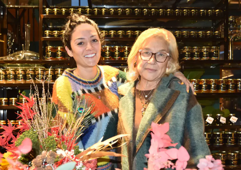 Smiling woman in colorful sweater hugs older woman in green coat amid shelves of jars and floral display in shop