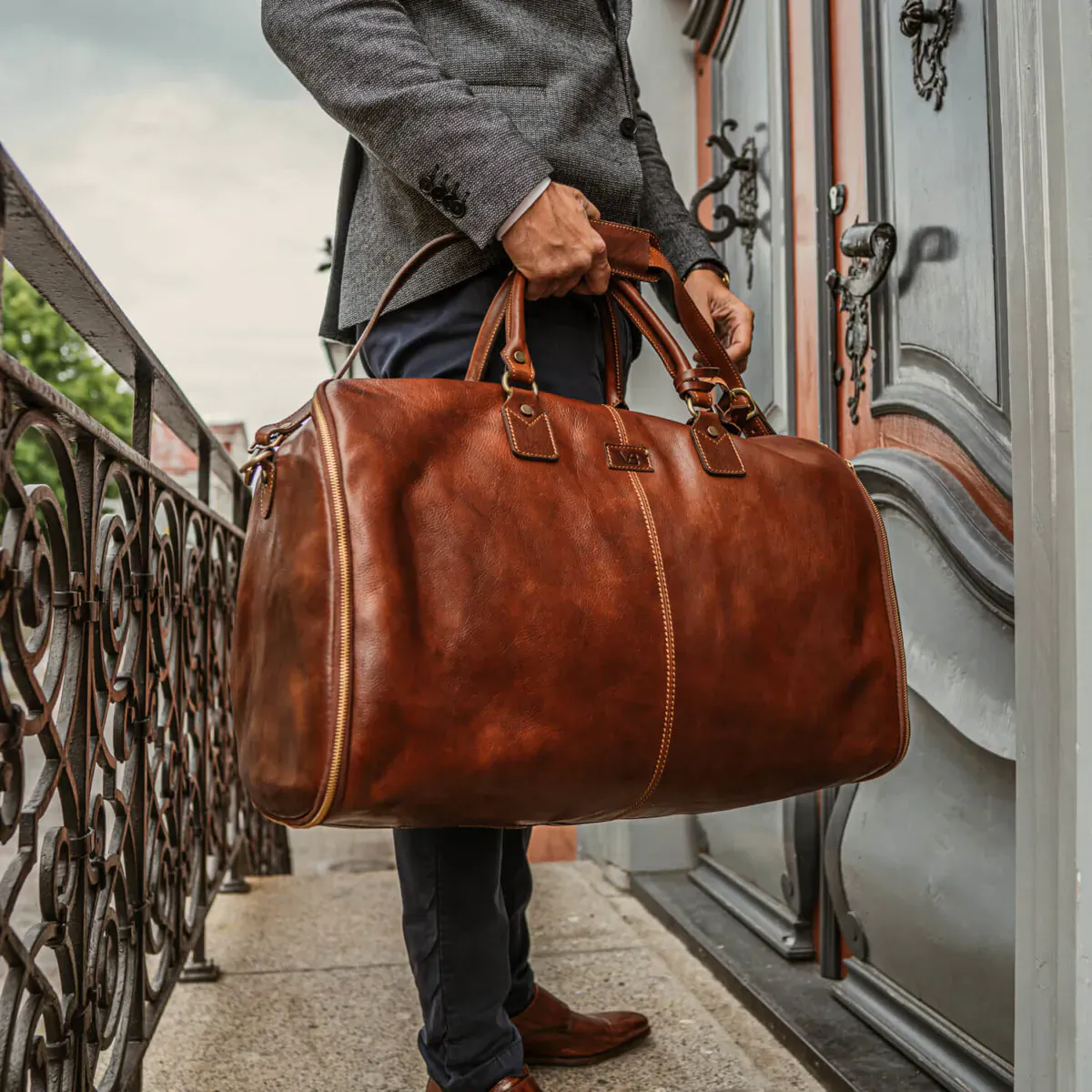 Man in gray suit holding tan leather duffel bag by ornate door on balcony railing