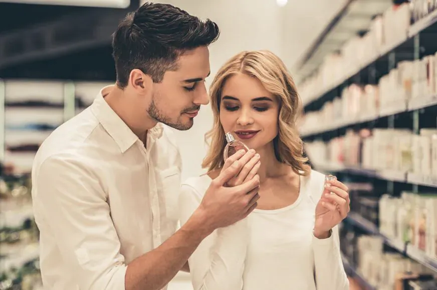 Young couple in store, man gently sniffing perfume sample held by smiling blonde woman amid shelves