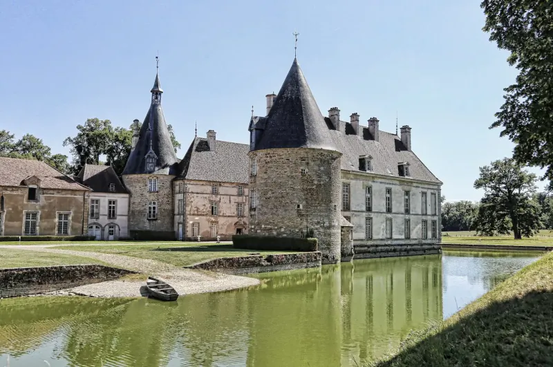 French château with slate turrets reflected in moat, surrounded by trees and lawns in Burgundy wine country.