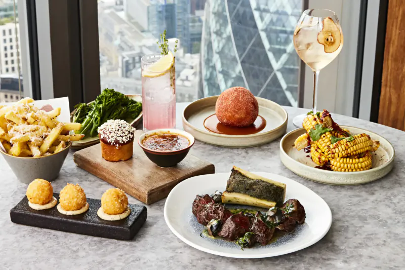 Duck and Waffle restaurant dishes on table: steak, red ball, fries, corn, croquettes, pink drink, wine glass, city skyline view.