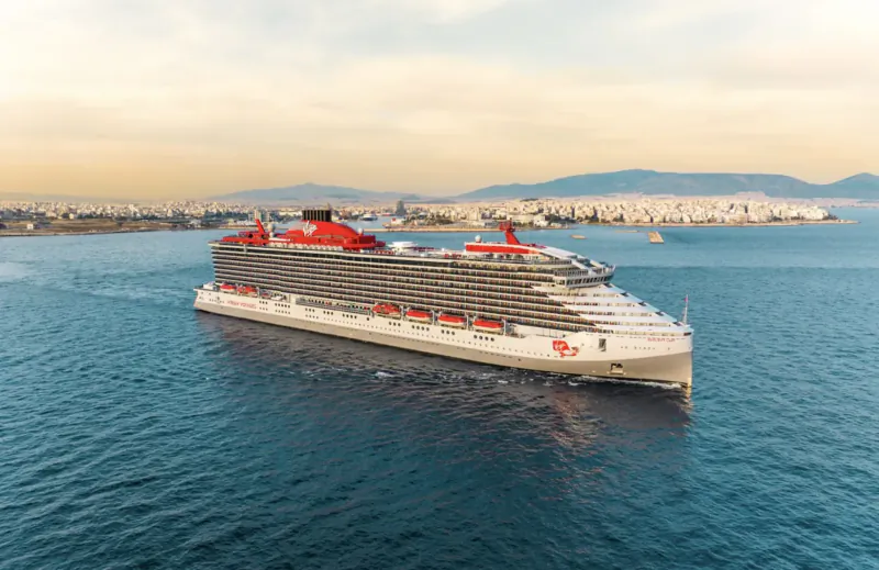 Large white Virgin Voyages cruise ship with red accents sailing on sea toward city and mountains at sunset