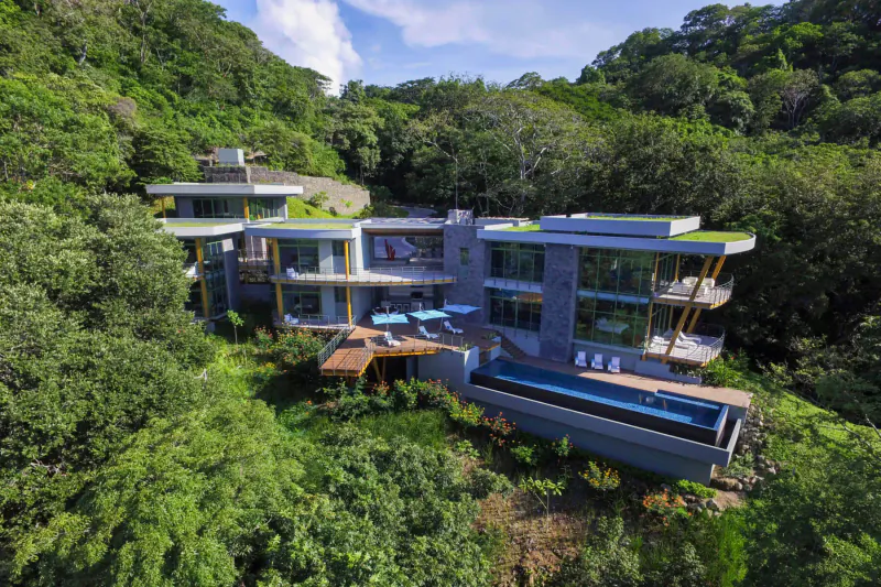 Modern luxury house with infinity pool and deck, perched on lush tropical hillside overlooking forest.
