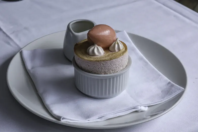 Ramekin of pink cremeux topped with whipped cream and chocolate orb, beside small white jug on white plate with napkin