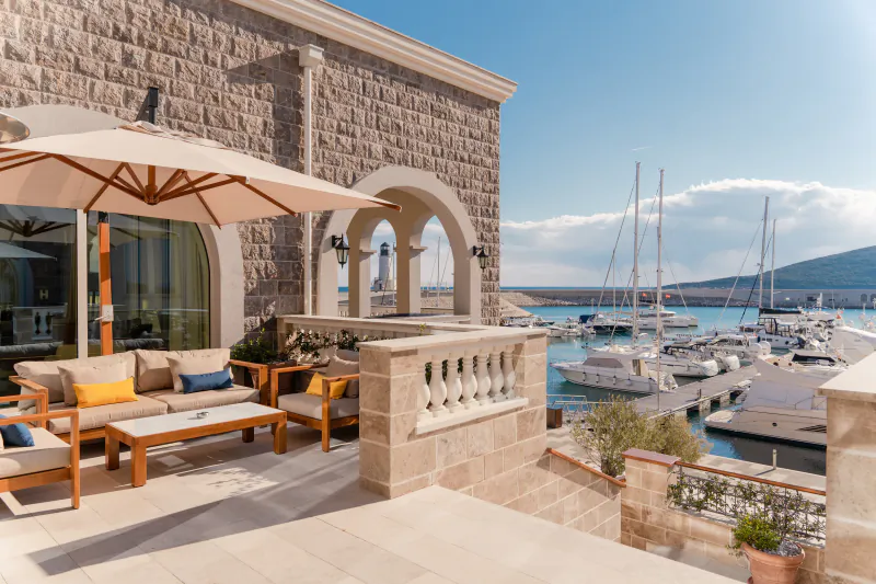 The Chedi Luštica Bay terrace with beige umbrella, yellow cushions on outdoor furniture, overlooking marina with yachts in Montenegro.