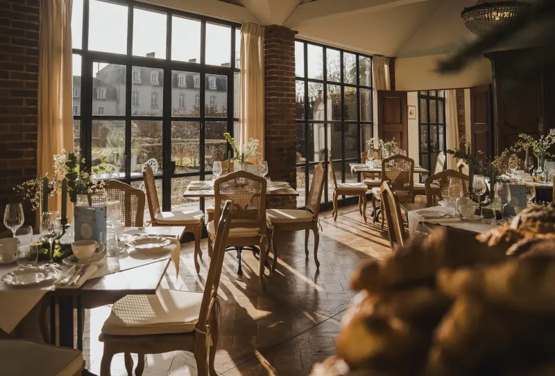 Elegant dining room at Le Jardin Privé with ornate chairs, tables set with glassware, fresh flowers, and crusty bread, large windows overlooking gardens.