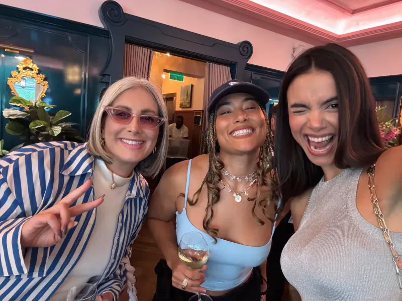 Three smiling women – one in striped shirt making peace sign, one in blue top with champagne, one in gray top – posing in gallery.