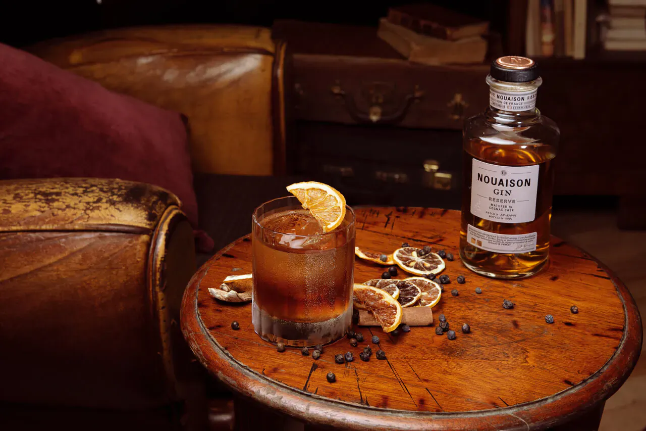 Hendrick's Gin Christmas cocktail in rocks glass with orange slice, dried peels, and juniper berries on wooden table beside leather chairs.