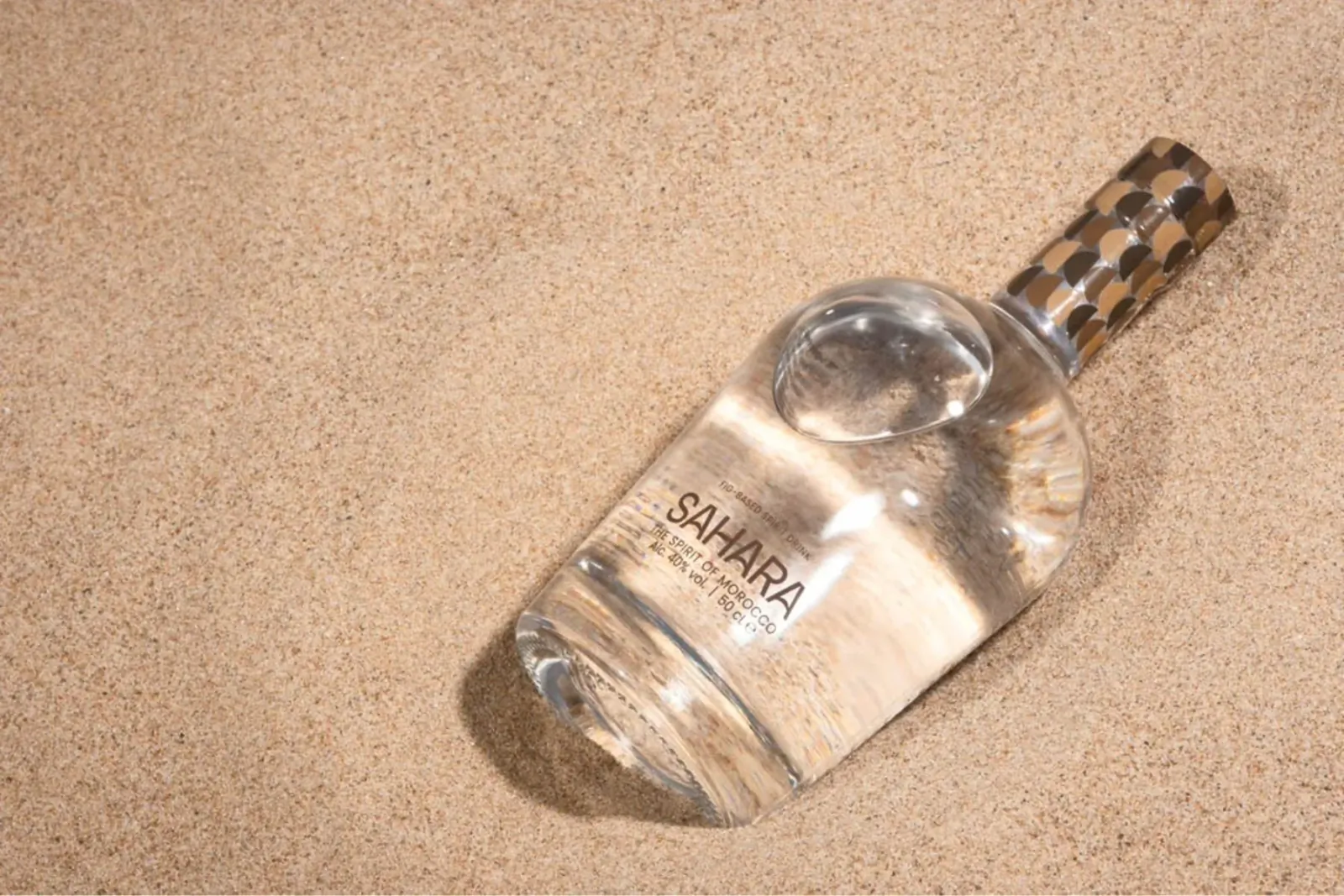 Sahara perfume bottle with clear liquid and gold cap on beige sand surface