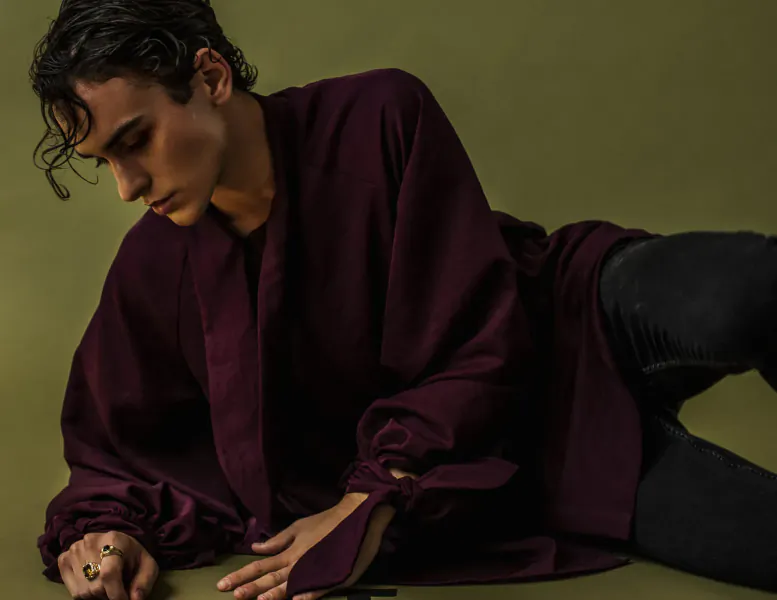 Javi Costa Polo reclines on green background in purple kimono-style robe and black pants, hand extended elegantly.
