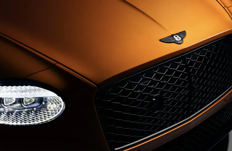 Close-up of orange Bentley Continental GT front: illuminated headlights, black grille, Bentley wings emblem.