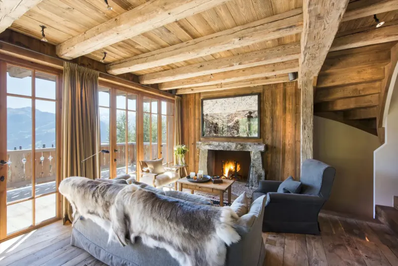 Luxury ski chalet living room with wooden beams, stone fireplace, fur-draped sofa, armchairs, and panoramic mountain views through large windows.