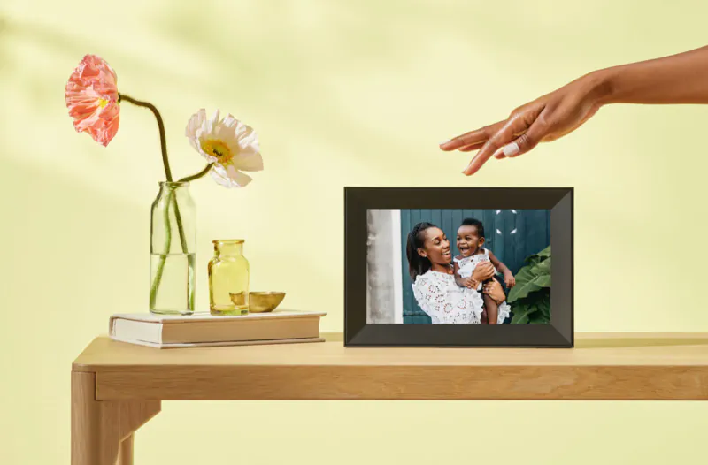 Aura smart photo frame on wooden table with flowers, displaying smiling mother holding baby boy.