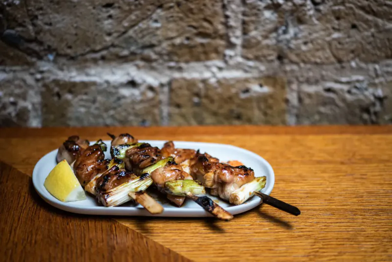 Grilled chicken and spring onion skewers on white plate with lemon wedge, on wooden table against brick wall.