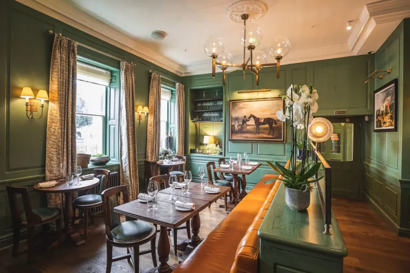 Elegant green dining room in Hotel Indigo Bath with wooden tables, chairs, chandelier, orchids, and landscape painting.