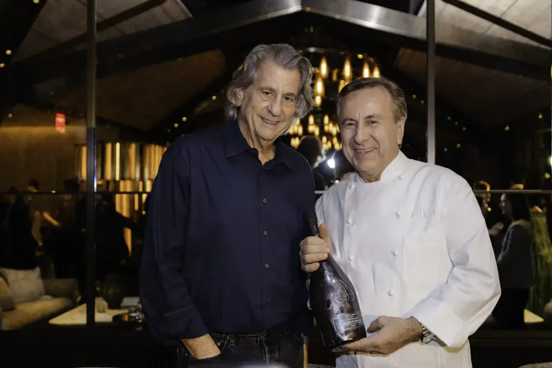 Two men smiling in modern NYC gallery: one in dark shirt, chef in whites holding Armand de Brignac bottle